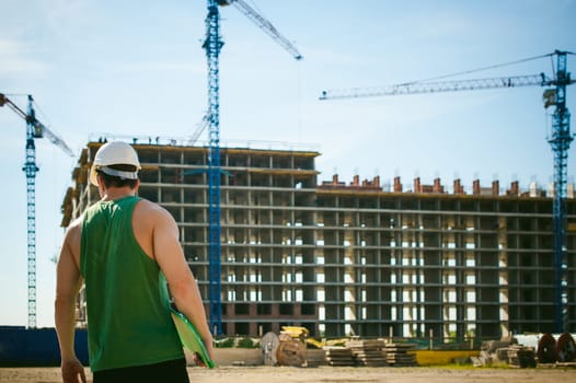 Young sexy men bodybuilder athlete. Manager engineer builder in white protective helmet stands on background of construction of building and construction cranes with documentation in hand
