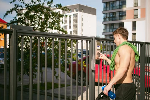 Handsome sexy male athlete man with naked torso, stands near gate, calling intercom to open door, after morning run in open air in urban courtyards, an early sunny summer morning