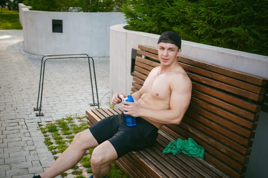 Handsome sexy male athlete man with naked torso resting on a bench in the courtyard of the house, after training drinks water from a bottle, an early sunny summer morning