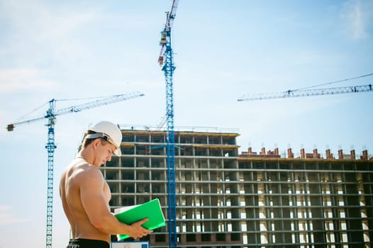 Young sexy male bodybuilder athlete with a bare torso. Manager engineer builder in white protective helmet stands on background of construction of building and construction cranes with documentation