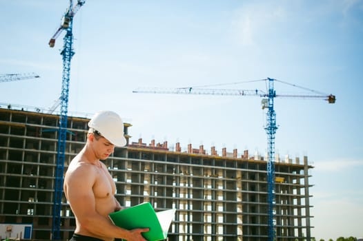 Young sexy male bodybuilder athlete with a bare torso. Manager engineer builder in white protective helmet stands on background of construction of building and construction cranes with documentation