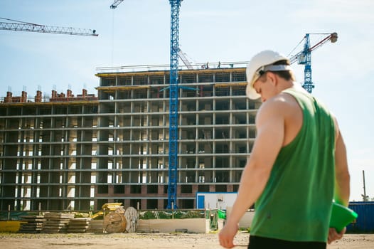 Young sexy men bodybuilder athlete. Manager engineer builder in white protective helmet stands on background of construction of building and construction cranes with documentation in hand