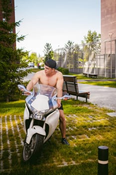 Handsome sexy male athlete man with naked torso in yard of house in parking lot sitting on a white sport motorcycle, an early sunny summer morning