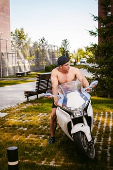 Handsome sexy male athlete man with naked torso in yard of house in parking lot sitting on a white sport motorcycle, an early sunny summer morning