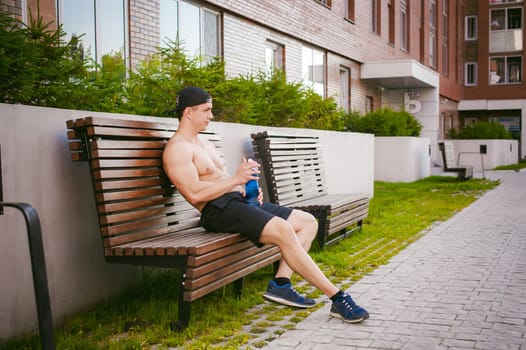 Handsome sexy male athlete man with naked torso resting on a bench in the courtyard of the house, after training drinks water from a bottle, an early sunny summer morning