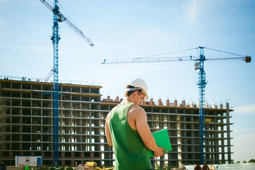 Young sexy men bodybuilder athlete. Manager engineer builder in white protective helmet stands on background of construction of building and construction cranes with documentation in hand