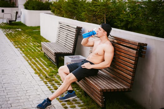 Handsome sexy male athlete man with naked torso resting on a bench in the courtyard of the house, after training drinks water from a bottle, an early sunny summer morning