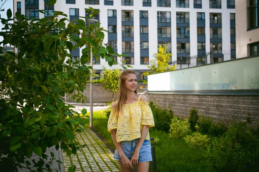 Young beautiful woman is strolling outdoors in the yard, against the backdrop of greenery and houses, warm summer sunny day