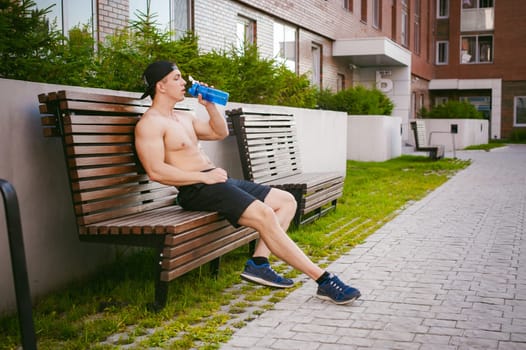 Handsome sexy male athlete man with naked torso resting on a bench in the courtyard of the house, after training drinks water from a bottle, an early sunny summer morning