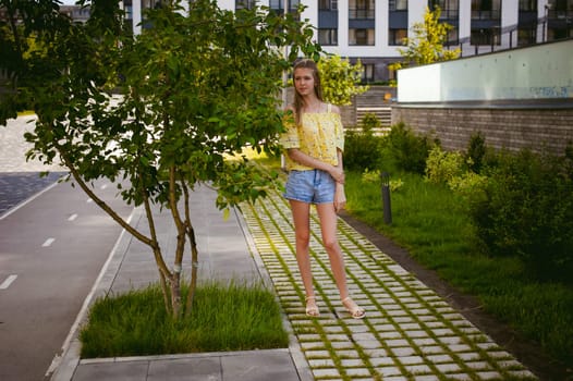 Young beautiful woman is strolling outdoors in the yard, against the backdrop of greenery and houses, warm summer sunny day