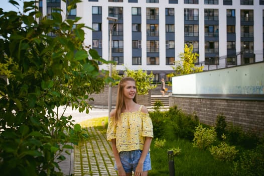 Young beautiful woman is strolling outdoors in the yard, against the backdrop of greenery and houses, warm summer sunny day