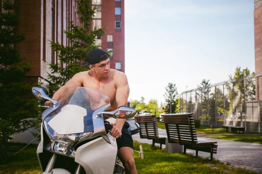 Handsome sexy male athlete man with naked torso in yard of house in parking lot sitting on a white sport motorcycle, an early sunny summer morning