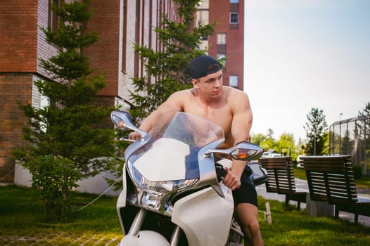 Handsome sexy male athlete man with naked torso in yard of house in parking lot sitting on a white sport motorcycle, an early sunny summer morning