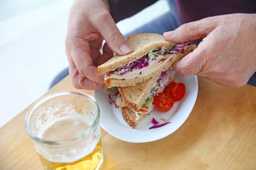 A man holds half a turkey sandwich on wheat bread with coleslaw, cherry tomatoes and a glass of beer