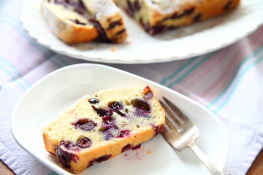 A serving of fresh blueberry cake on a white dish, rest of cake in background