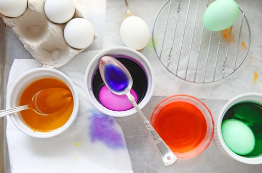 Various bowls with dye, eggs being dyed, spoons, a small rack for drying with dye stains on paper towels underneath