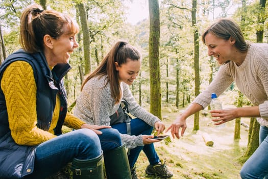 Teenager is using her smart phone while taking a break from a hike with her mother and grandmother. They are all looking at the phone together.