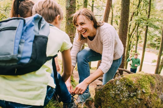 Happy family are hiking through the woodlands together. The mother has stopped to help her son tie the laces on his hiking boots.