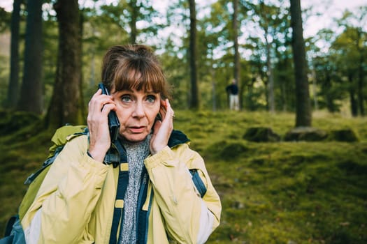 Senior couple are lost in the woods. The woman is trying to get signal on her phone and her partner can be seen in the background.