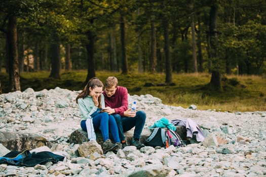 Two teenage siblings are sitting on a pebble beach after hiking. They are both looking at the girls' smart phone.