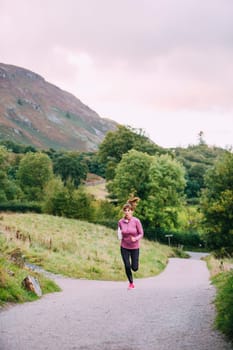 Senior woman is running along a trac path in the Lake District with a water bottle.