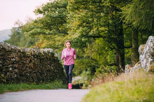 Senior woman is running along a trac path in the Lake District with a water bottle.