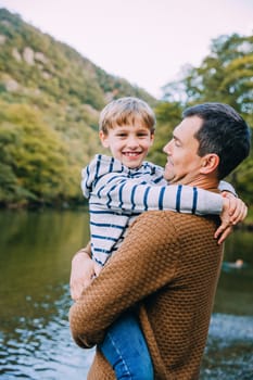 Little boy is smiling for the camera while being held by his father. They are standing near a lake.