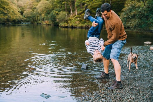Little boy and his father have been hiking and found a pond. His father is holding the boy upside down, teasing to dunk him in the water.