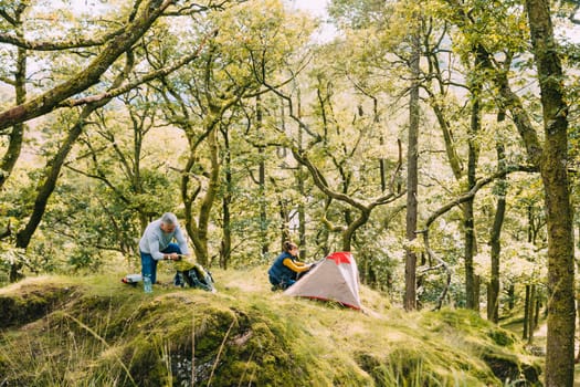 Senior couple have chosen their spot in the woodlands to set up for wild camping. The woman is setting up the tent and the man is getting things from a backpack.