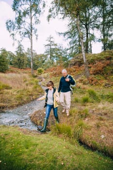 Senior couple are enjoying a walk through the woodlands together. They have come across a small stream which they are stepping over.