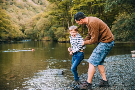 Little boy and his father have been hiking and found a pond. His father is holding the boy, teasing to throw him in the water.