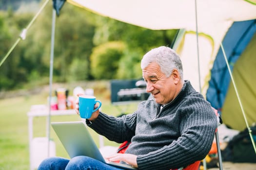 Senior man is relaxing in a chair by his tent in the campsite. He is enjoying a cup of tea while using a laptop.