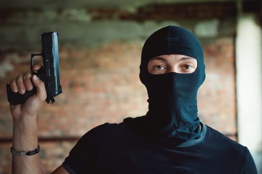 Athletic man in a balaclava, ski mask on his head, holds a pistol in his hand, lifting up a barrel, a dangerous gangster