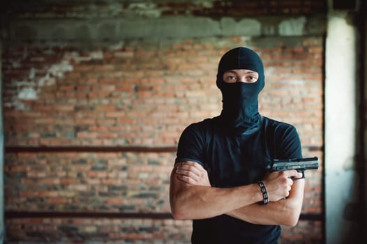 Athletic man in a balaclava, ski mask on his head, holds a gun in his hand, with his arms crossed over his chest