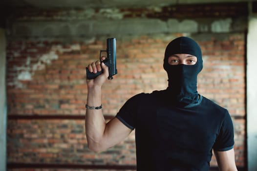 Athletic man in a balaclava, ski mask on his head, holds a pistol in his hand, lifting up a barrel, a dangerous gangster