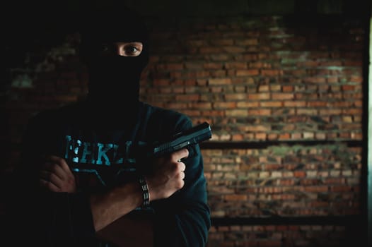 Athletic man in a balaclava, ski mask on his head, holds a gun in his hand, with his arms crossed over his chest