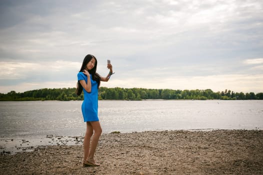 Young attractive Asian woman dressed in a blue dress, with long dark hair. Makes emotional selfie portrait photo on smartphone, on background of pond river outdoors by summer day