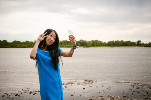 Young attractive Asian woman dressed in a blue dress, with long dark hair. Makes emotional selfie portrait photo on smartphone, on background of pond river outdoors by summer day
