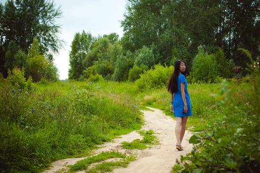 Young attractive Asian woman dressed in a blue dress, with long dark hair. Walks outdoors on a dirt road in nature among the greenery