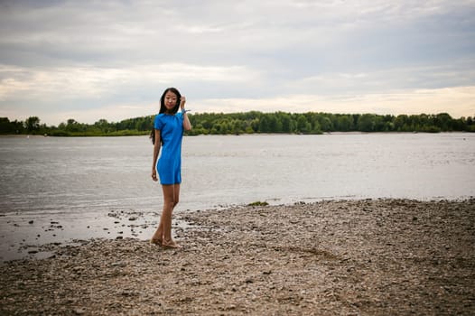 Young attractive Asian woman dressed in a blue dress, with long dark hair. Emotional portrait photo on background of pond river outdoors by summer day