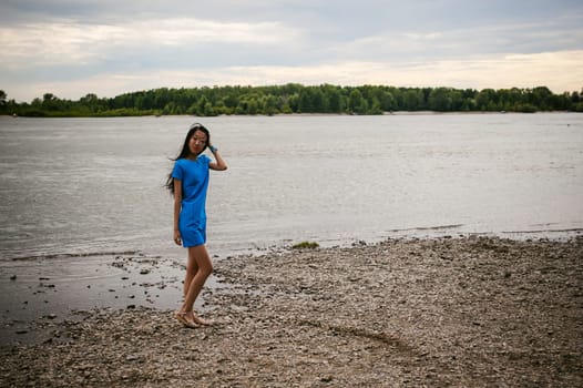 Young attractive Asian woman dressed in a blue dress, with long dark hair. Emotional portrait photo on background of pond river outdoors by summer day