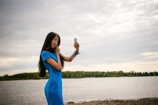 Young attractive Asian woman dressed in a blue dress, with long dark hair. Makes emotional selfie portrait photo on smartphone, on background of pond river outdoors by summer day
