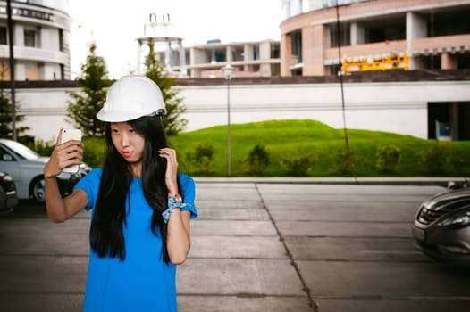 Asian woman in a blue dress and white protective helmet. engineer supervises inspection and control of works at construction site. Makes selfi on smartphone