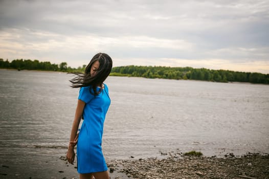 Young attractive Asian woman dressed in a blue dress, with long dark hair. Emotional portrait photo on background of pond river outdoors by summer day