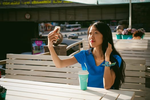 Cute asian young woman in summer cafe outdoors. girl In a blue dress, with long hair in simple light cozy interior of restaurant Urban style, Communicates on a cell phone holding it in his hands