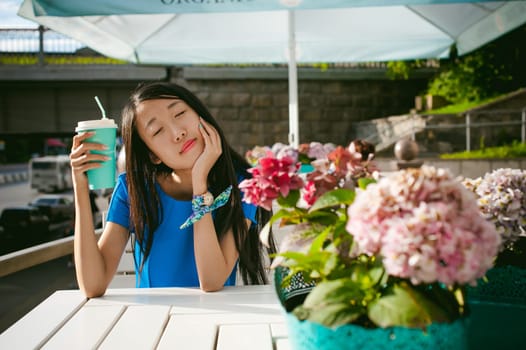 Cute asian young woman in summer cafe outdoors. girl In a blue dress, with long hair in simple light cozy interior of restaurant Urban style, drinking coffee drink, sitting at table with flowers
