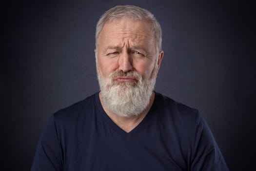A 60 year old man with gray beard posing spoiled grimace in the studio