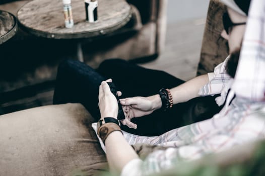 young man with beard in light checkered jacket with hood in a cap and sunglasses  sitting on a sofa in a summer cafe uses a smartphone