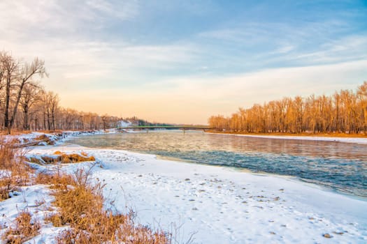 Landscape of the Bow River with snow covered banks, warm light and a bridge in the distance.