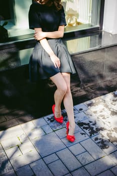 Beautiful woman in a dark stylish dress strolls along the street, near boutiques. Slender legs in red shoes